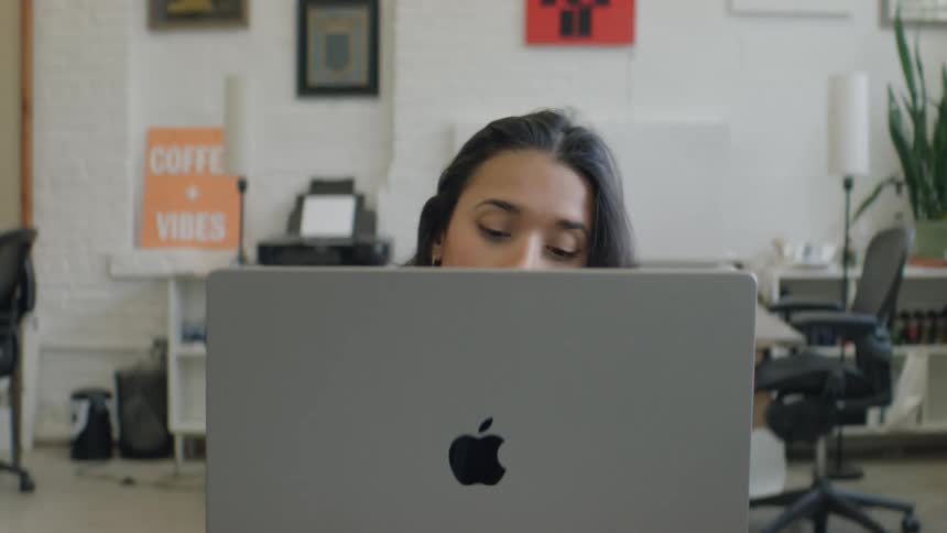 Woman working on a laptop in an office with desks chairs and decorative frames in the background