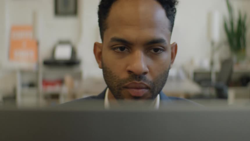 A young black man sits in an office working on his laptop as he types on the keyboard