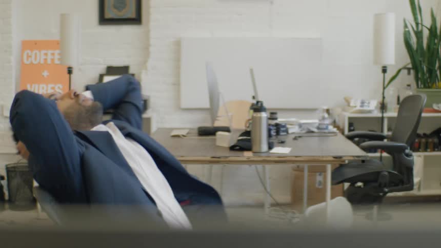 Man leaning back in a chair with hands behind his head in an office with desks chairs and work items in the background