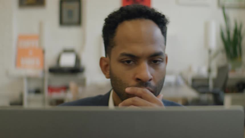A man with a serious expression sits behind a laptop in a bright office resting his chin on his hand while looking at the screen before shifting his gaze downward and back up as he continues to work amidst desks and wall decorations