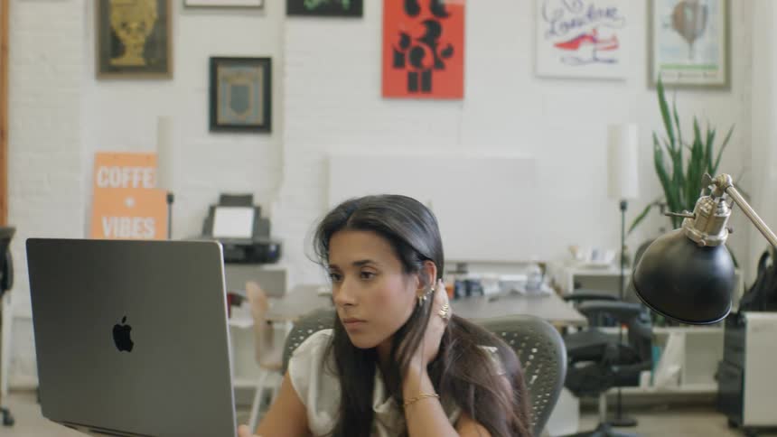 A young woman is focused on her work at a laptop in a bright modern office spaceshe begins by adjusting her hair behind her ear while looking at the screen then settles into a steady rhythm of typing