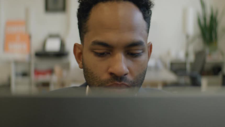 A man with a serious expression sits in an office and looks intently at a laptop screen while working with a softfocus background showing typical workspace elements like plants and furniture
