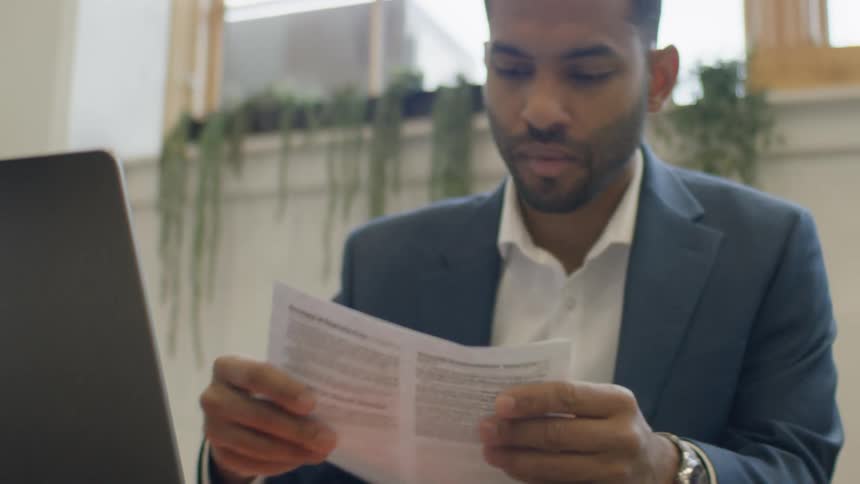 Man in a suit reading a printed document while working at a desk with a laptop indoors
