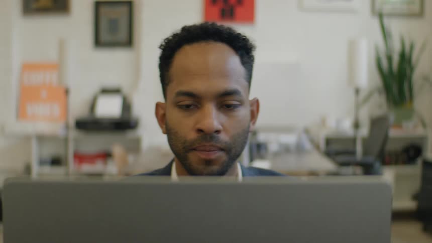 A young black man sits behind a desk in an office at midday wearing a blue blazer suit as he works on his laptop