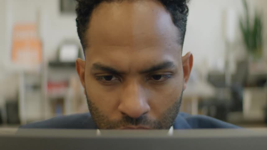 A young black man sits behind a desk in an office at midday wearing a blazer suit and typing on his laptop as daylight fills the room