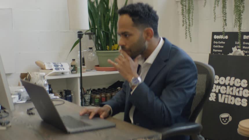 Man working on a laptop seated at an office desk with computers plants and an information board in the background