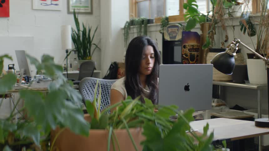 A young woman sits behind a desk in an office at midday typing on her laptop the room is bright and filled with potted plants giving the workspace a calm natural feel as she works