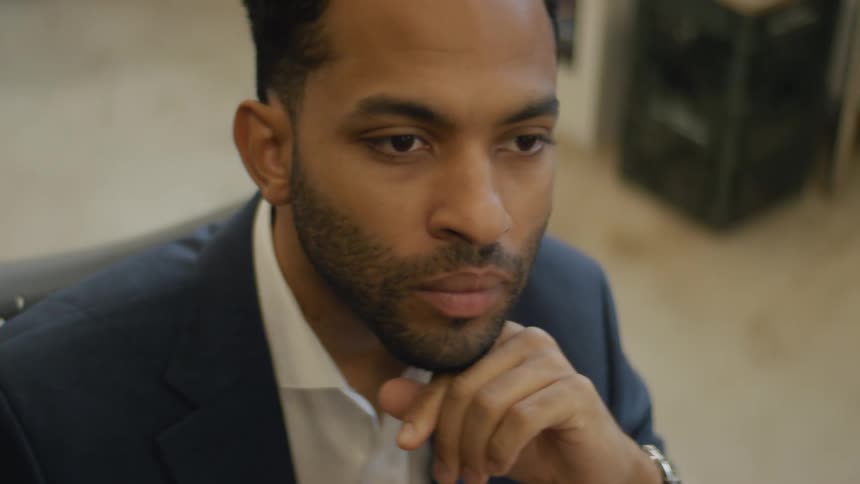 Young adult black man sitting down with his hand on his chin and a contemplative facial expression indoors