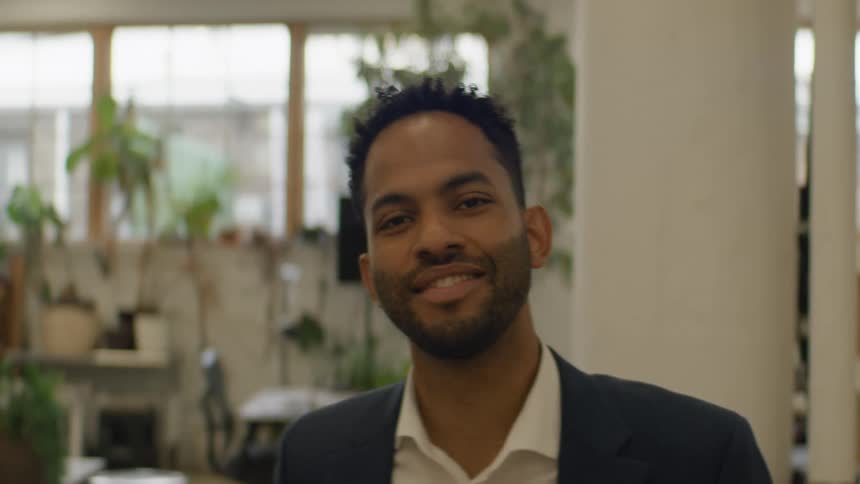 A man in a navy suit and white shirt looks forward with a calm and professional mood in a bright office filled with plants