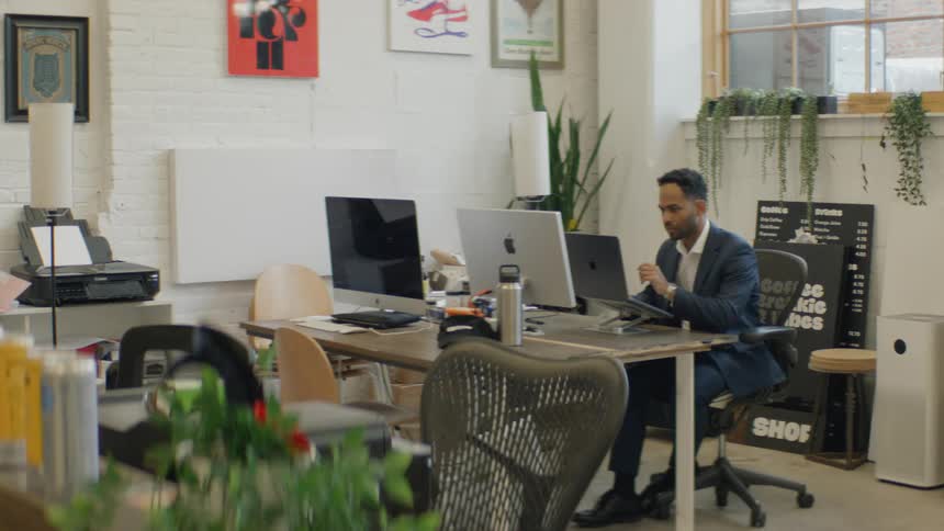 A man in a navy suit and white shirt sits at a long wooden desk using a laptop in a bright office filled with posters and plants looking forward with quiet concentration