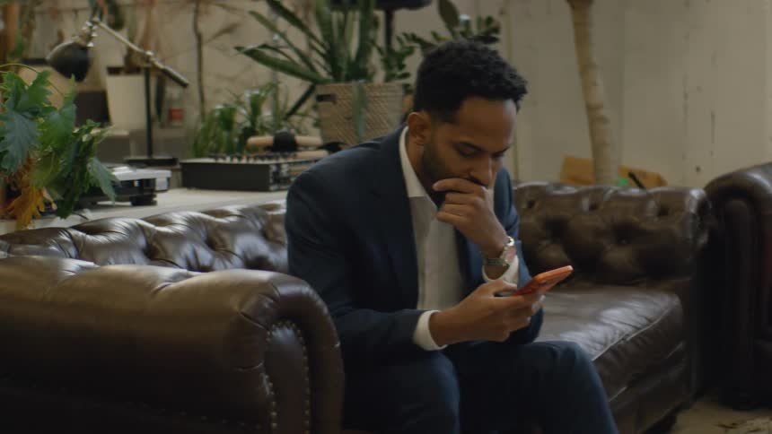 A young man sits on a couch at midday wearing a blue blazer and scrolling through his orange iphone 17