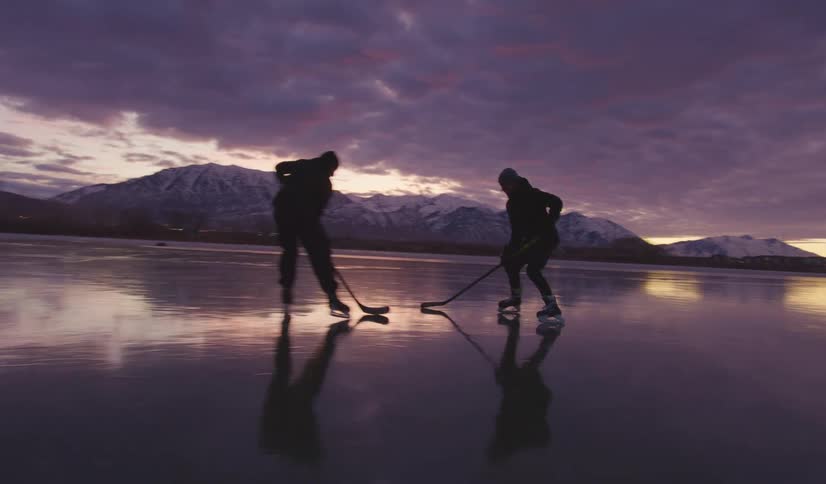 People playing ice hockey on a frozen lake at dusk with mountains in the background