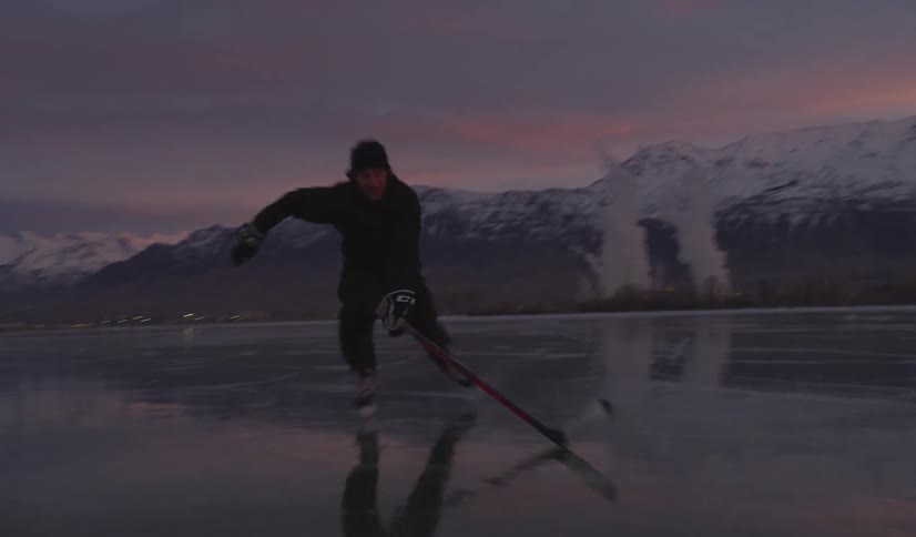 Adult man ice skating while carrying a hockey stick in his hand over a frozen river surrounded by mountains outdoors during dusk or dawn