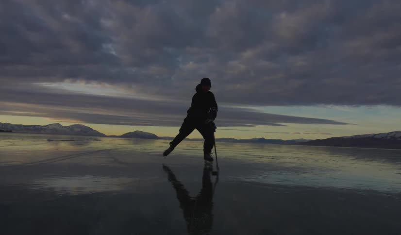 Adult man skating on ice and playing pond hockey over a frozen body of water with mountains on the background during dusk or dawn
