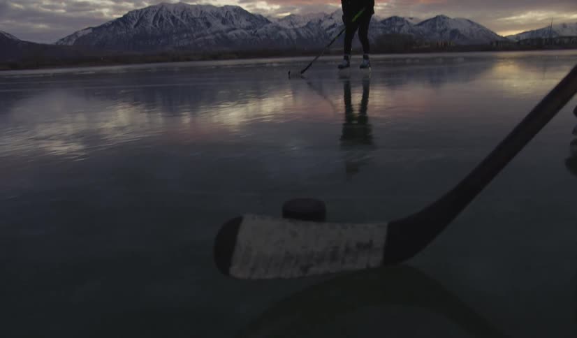 Two people are playing ice hockey on a frozen lake at sunset the sky is orange and you can see tall mountains far away with snow on the top