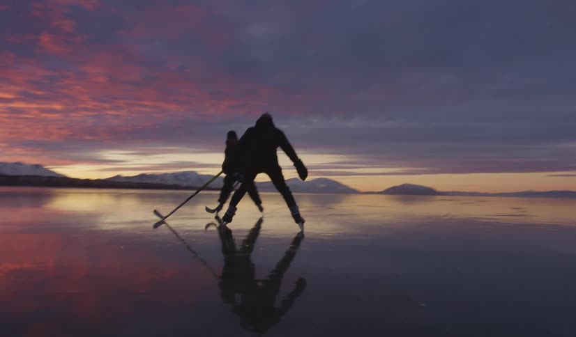Two young men playing ice hockey with mountains along the frozen coast