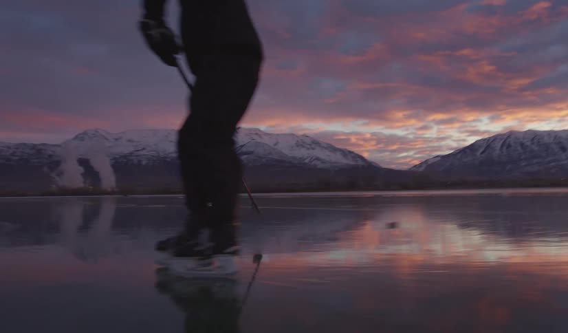 Two people play ice hockey on a vast frozen lake during a vibrant pink sunset