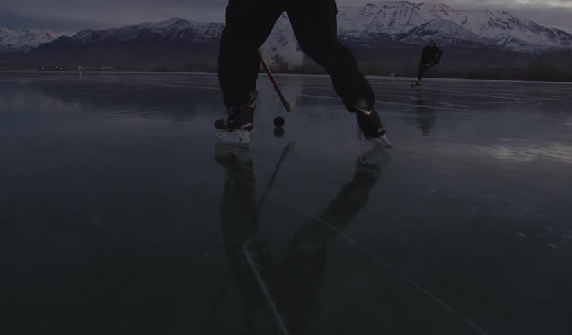 Two men play hockey on a vast frozen lake at dusk and the camera follows their skates and sticks as they move across the reflective ice with snow capped mountains and a cloudy sunset