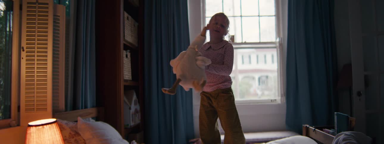 A young girl stands on a bed holding a large stuffed goose in a dimly lit room with blue curtains looking forward with a playful mood
