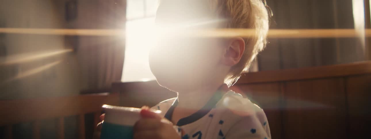 A young boy with blonde hair wearing starpatterned pajamas drinks from a blue sippy cup in a sunlit room