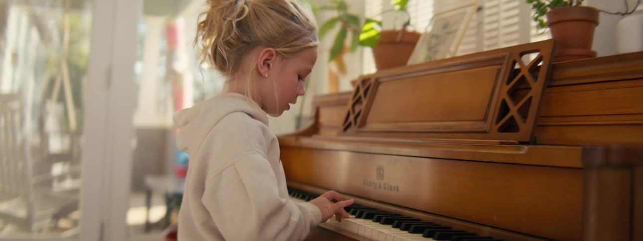 A young girl sits in a living room playing a grand piano sunlight comes through the window and reflects softly across the room as she presses the keys