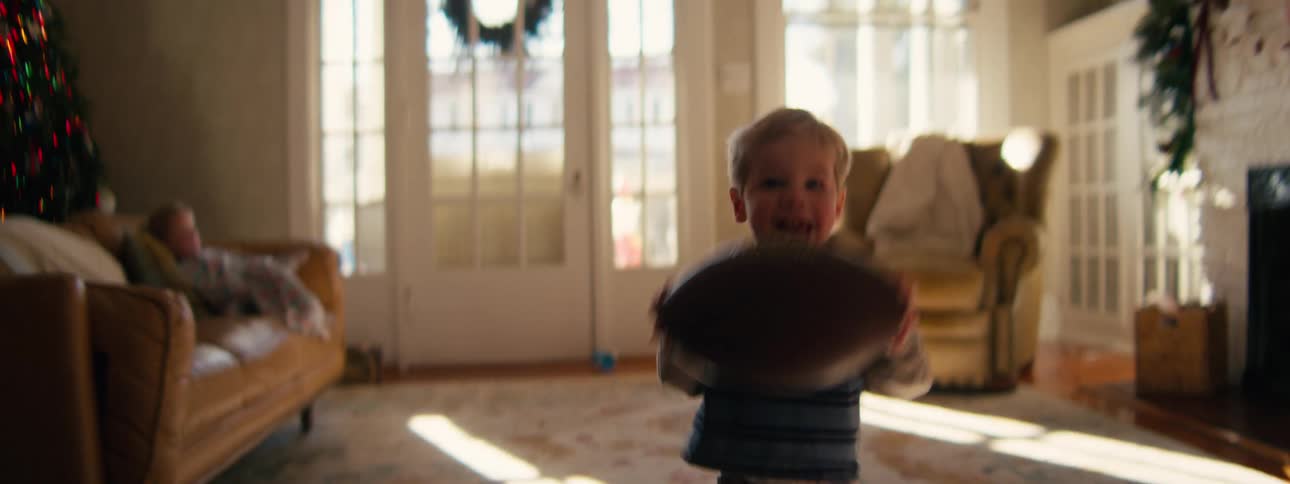 A young toddler in a striped shirt runs toward the camera through a sunlit living room while holding an american football