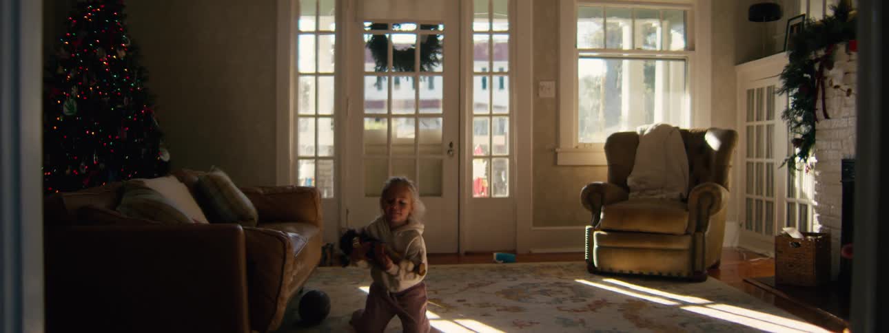 Child on the living room rug holding a toy with a christmas tree in the background and sunlight streaming through glass doors