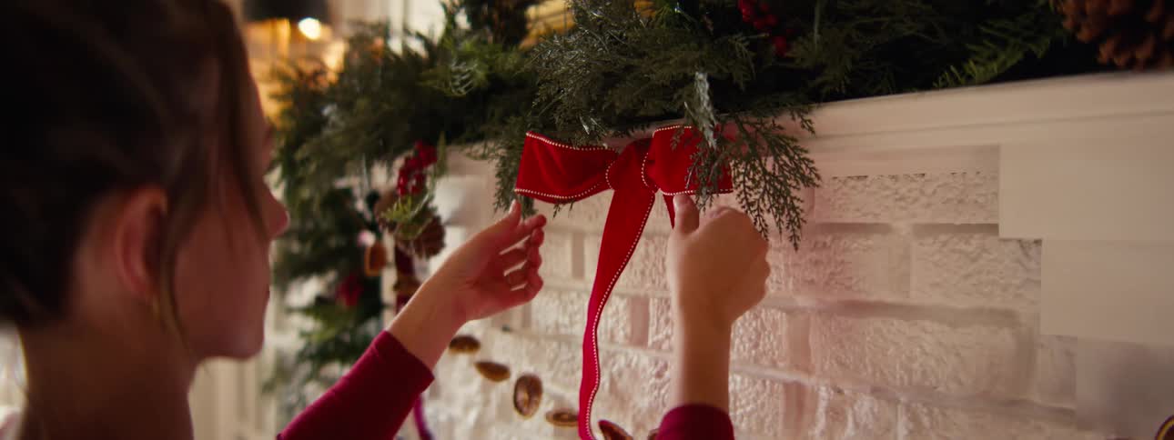 Woman adjusts a red bow tied to fir branches lying on the mantel