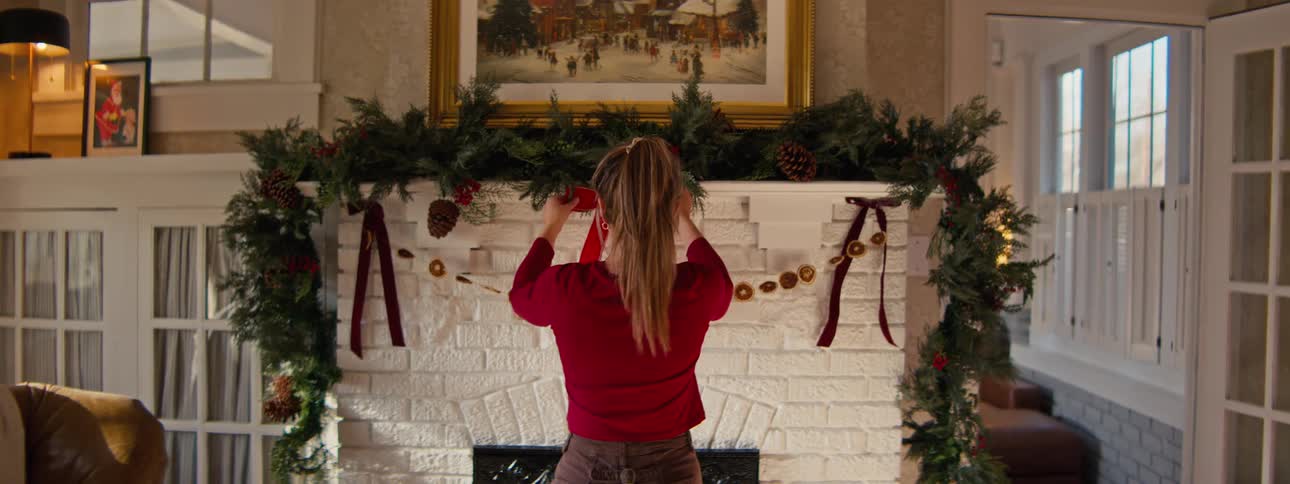 Woman adjusting garland and red ribbons over a fireplace decorated for christmas