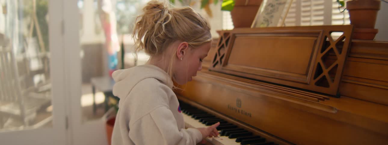 A young girl wearing a tan sweatshirt plays a piano in a bright room looking forward with a focused and joyful mood