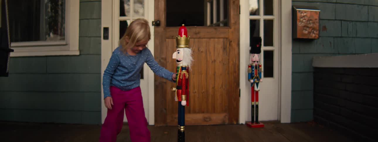 A young girl wearing a blue longsleeved shirt and pink pants plays with nutcracker decorations on a wooden porch looking forward with a playful mood
