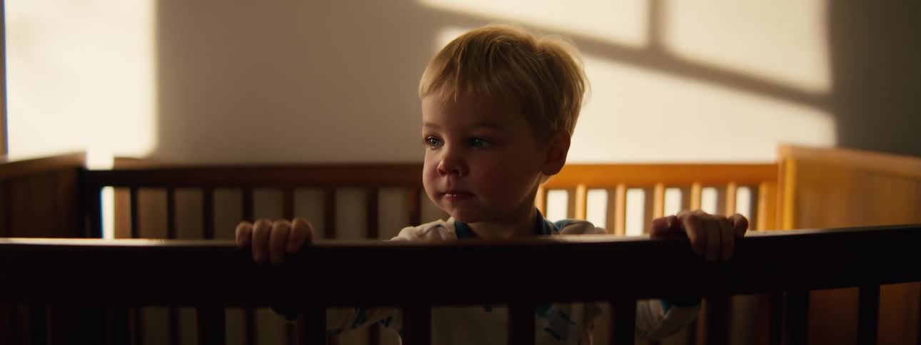 A toddler stands on his bed holding the wooden rails for support sunlight streams into the room lighting up his smiling face as he looks around happily