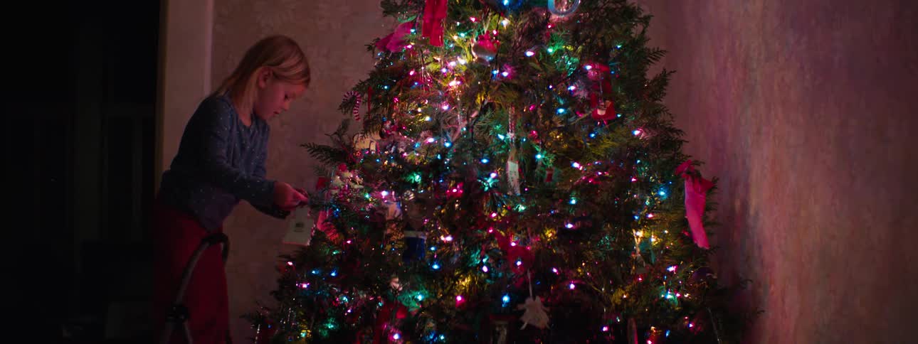A young girl hangs a decoration on a christmas light inside a room at night the lights glow softly beside the wall brightening the space as she reaches up to place the ornament