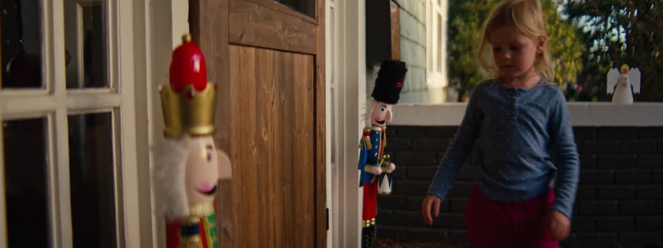 A young blonde girl in a blue shirt and pink pants carefully arranges large nutcracker decorations on a front porch before walking through the wooden front door of the house