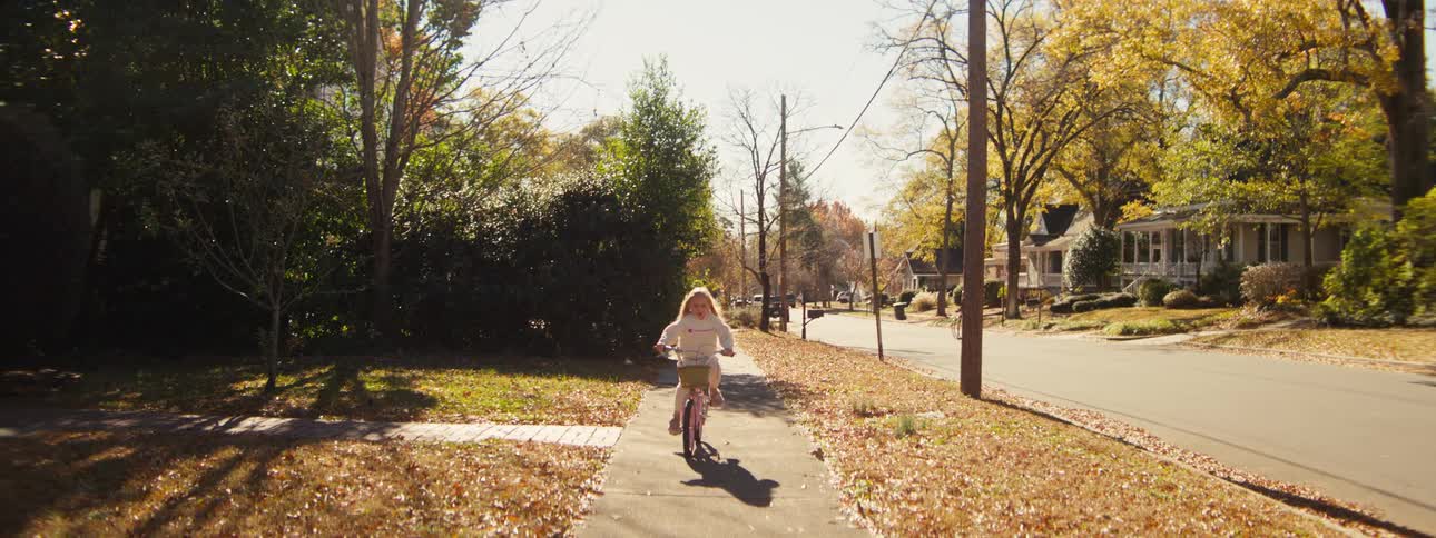 A young girl with long blonde hair rides a pink bicycle toward the camera along a sundrenched suburban sidewalk covered in autumn leaves passing by residential houses and mature trees under a bright clear sky
