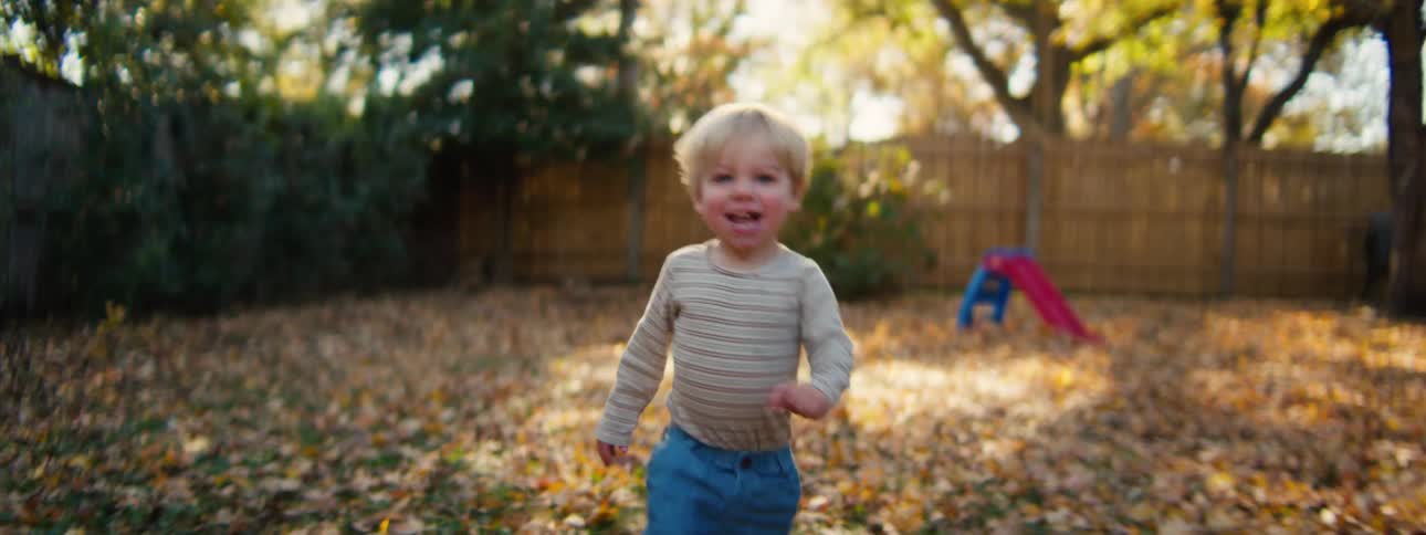Boy walks through a backyard covered with fallen leaves with a slide in the background