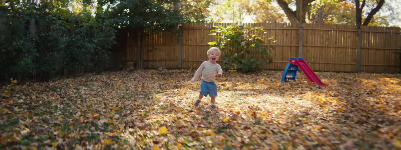 Child runs across a backyard covered with fallen leaves with a slide in the background