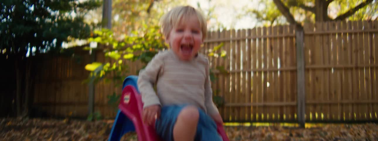 A young boy with blonde hair laughs and smiles with excitement as he slides down a small red and blue plastic slide into a pile of autumn leaves in a backyard