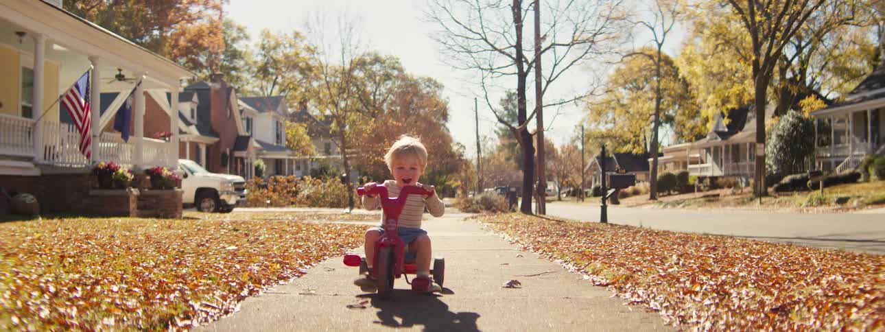 A young child wearing a tan sweatshirt rides a red tricycle down a leaf covered sidewalk looking forward with a joyful and energetic mood