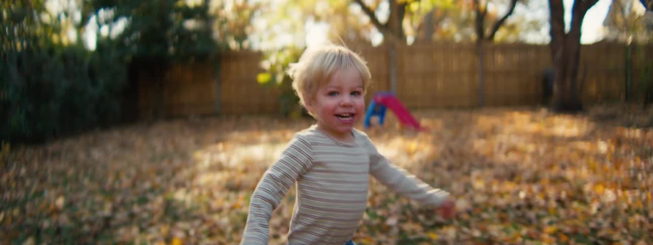 Child running in a backyard covered with dry leaves with a wooden fence and trees in the background