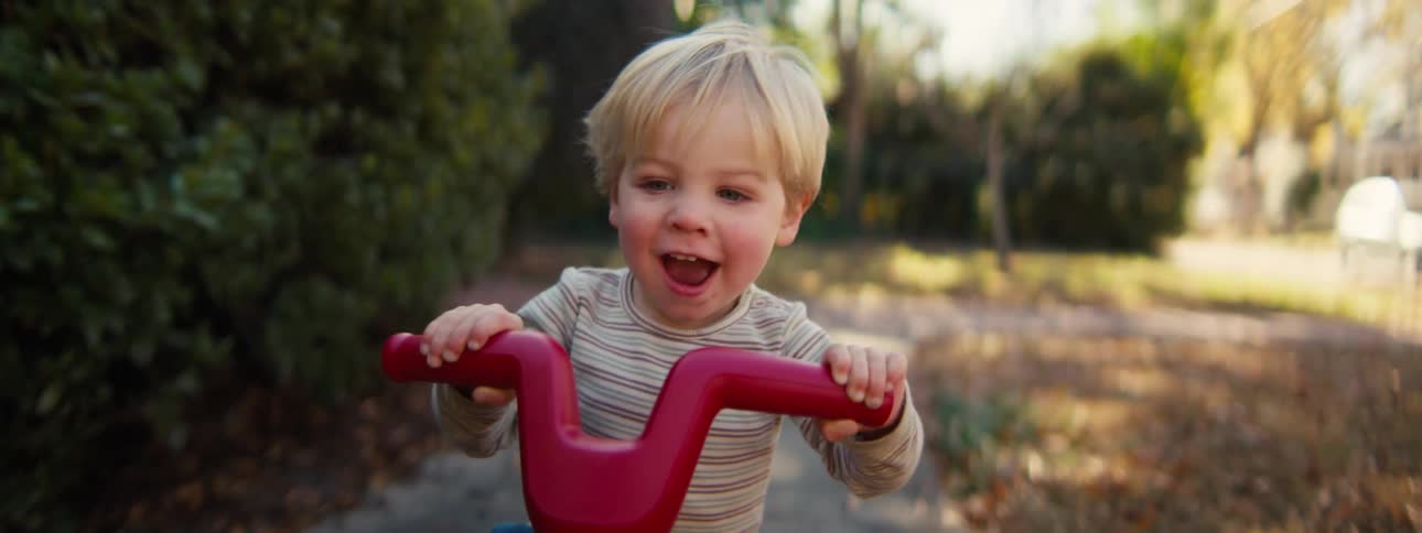 A toddler rides a small scooter outdoors on a sidewalk in a suburb setting