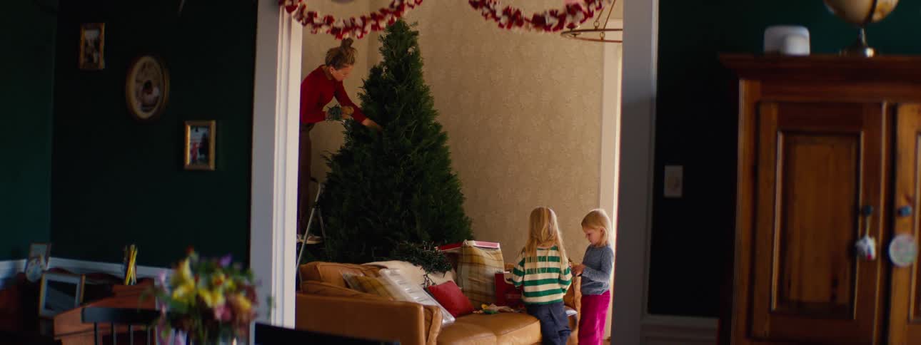 Children arranging christmas decorations in the living room while an adult sets up a tree in the background