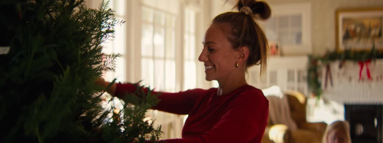 Woman smiles as she fixes the christmas tree with a string of lights on her arm as her girls play