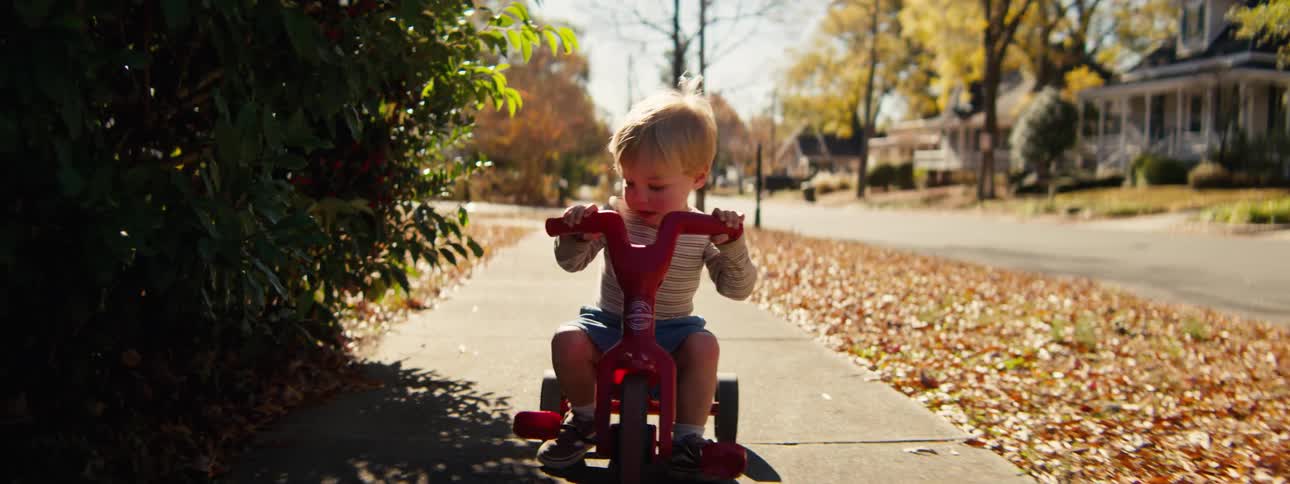 A happy toddler rides a red tricycle along a sidewalk covered in fallen autumn leaves