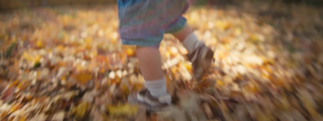 A young boy with blonde hair runs through a backyard filled with fallen autumn leaves toward a wooden fence