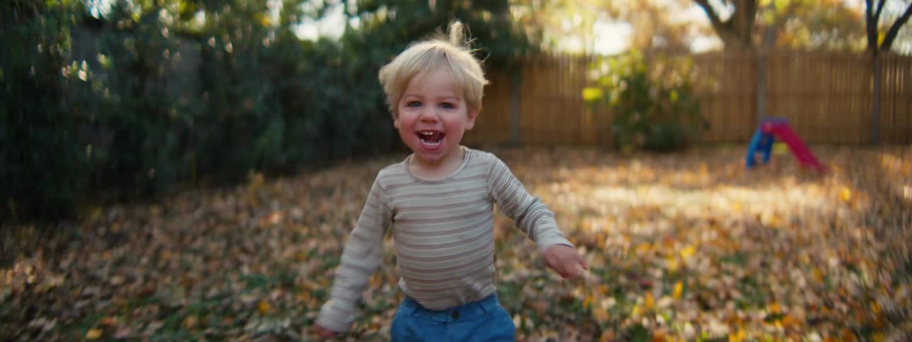 Boy runs through a backyard covered with fallen leaves with a childrens toy in the background