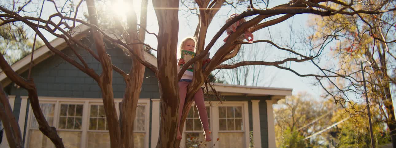 Child sitting on a tree branch reads a book in front of a residential house