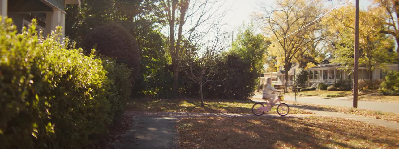 A young girl with long blonde hair rides a pink bicycle down a sunlit sidewalk and turns onto a residential street lined with autumn leaves and houses