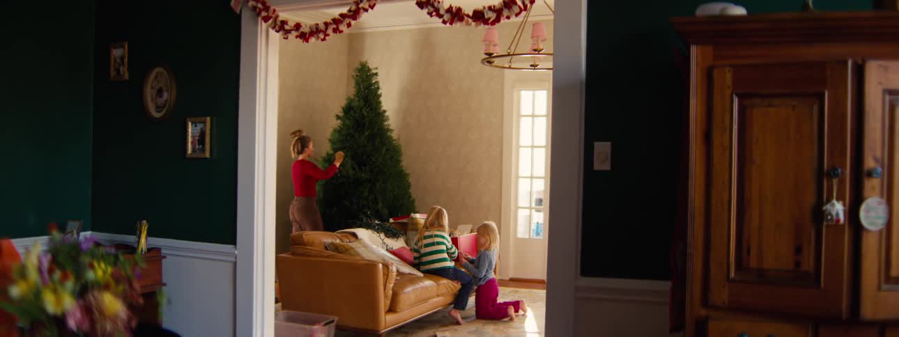A woman sets up a christmas tree while one girl sits on the sofa and the other kneels beside it
