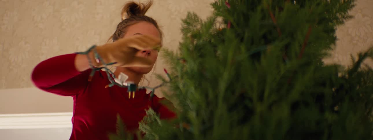 A woman is seen from the waist up close to a room ceiling hanging christmas lights on a tree near the top